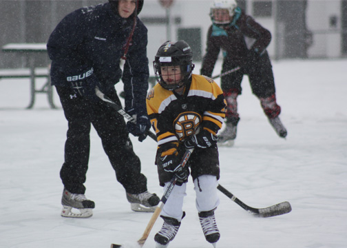 Photo of young boy wearing protective hockey helmet, holding hockey stick to the ice - 2 adults behind him are also holding hockey sticks to the ice 