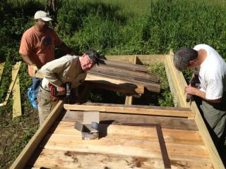 Photo of 3 men constructing wooden walkway, 2 of the 3 are bent over working on the walkway, one is standing back from it.